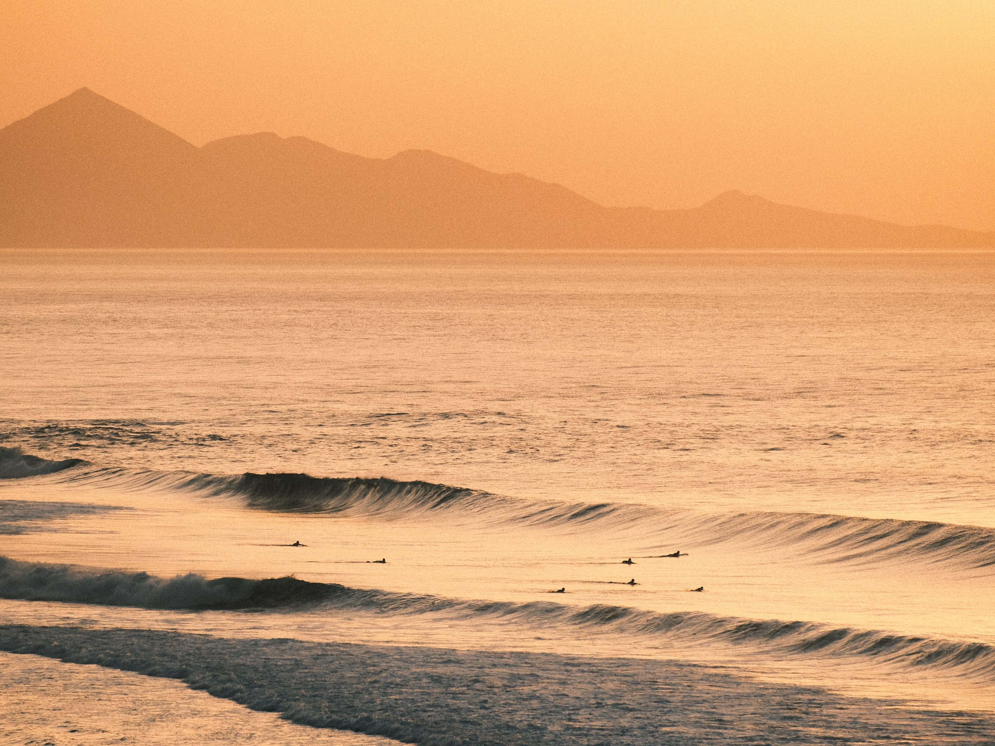 Surfers waiting in the line-up at golden hour with a mountain island on the horizon, Fuerteventura