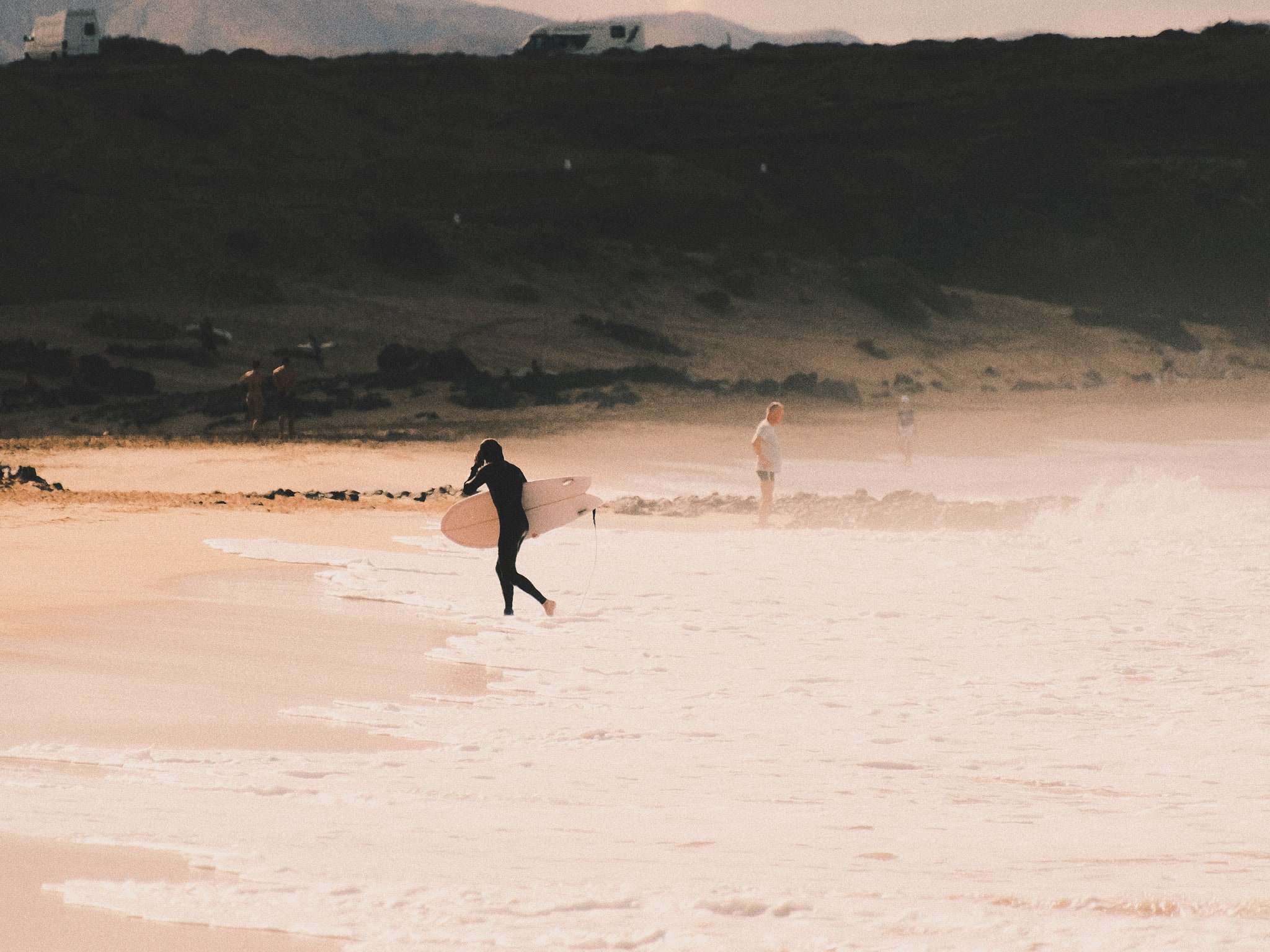 Surfer carrying a board into the sea on a sandy beach with volcanic rock behind